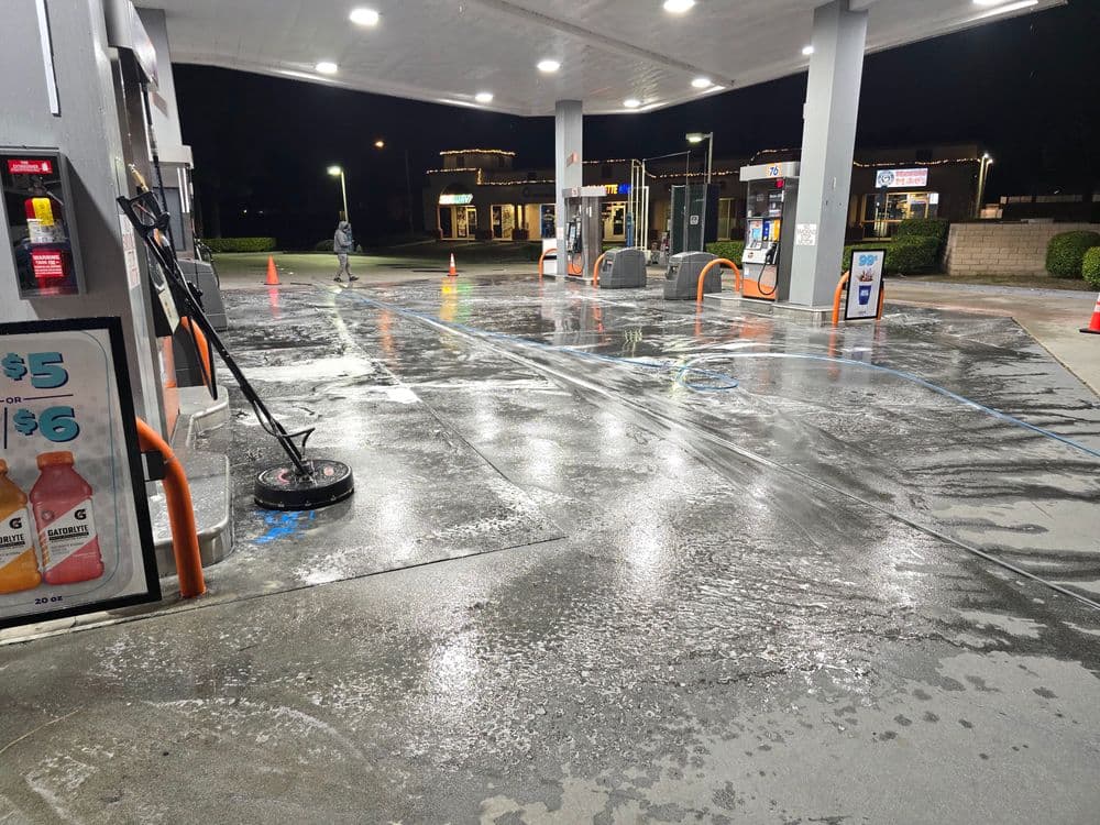 Nighttime gas station with wet concrete, cleaning equipment, and illuminated fuel pumps.