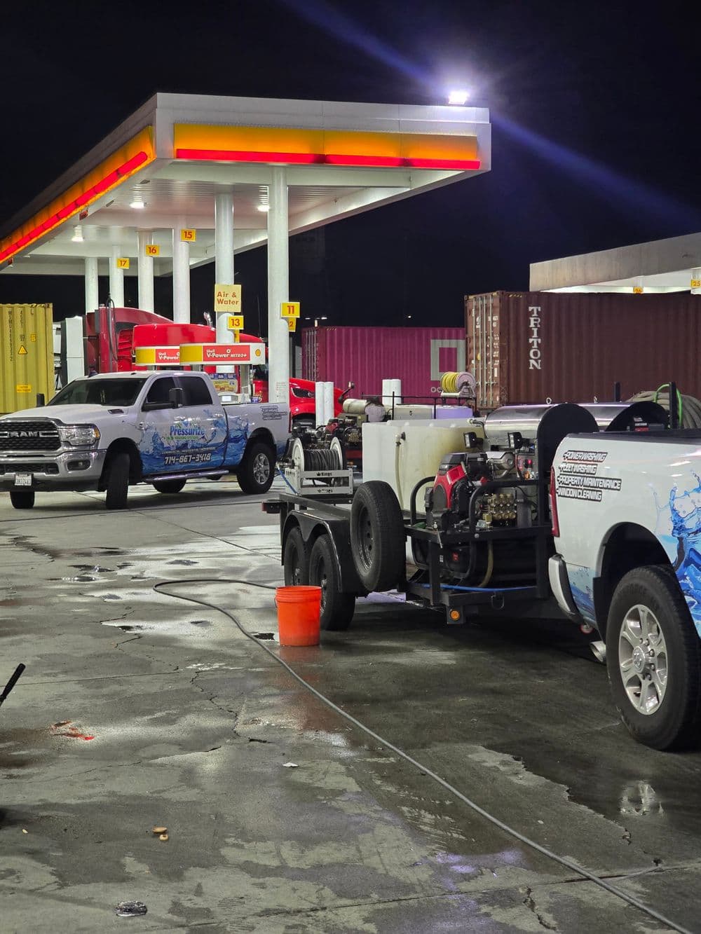 Pressure washing trucks at a gas station, nighttime with cargo containers in the background.